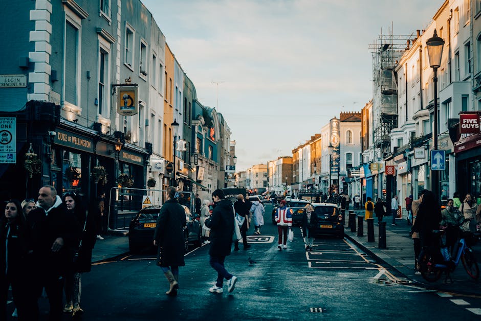 A street scene on Wanstead High Street showcasing a variety of commercial buildings, cafes, and pubs with retail signage visible. Pedestrians are walking along the sidewalk and crossing the street, so