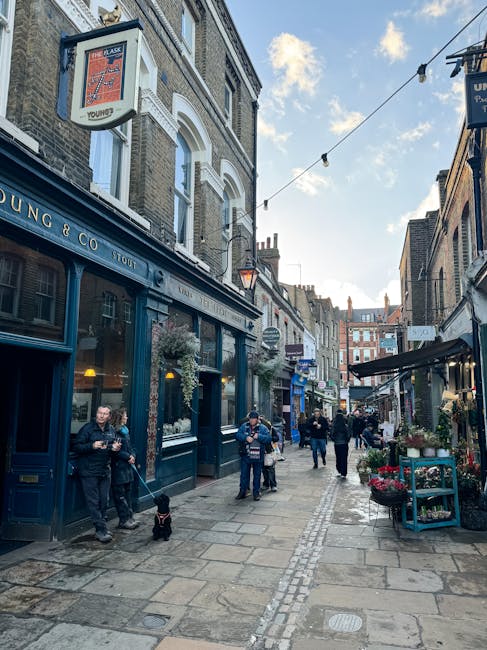 A street scene on Wanstead High Street showcasing a variety of commercial buildings, cafes, and pubs with retail signage visible. Pedestrians are walking along the sidewalk and crossing the street, so